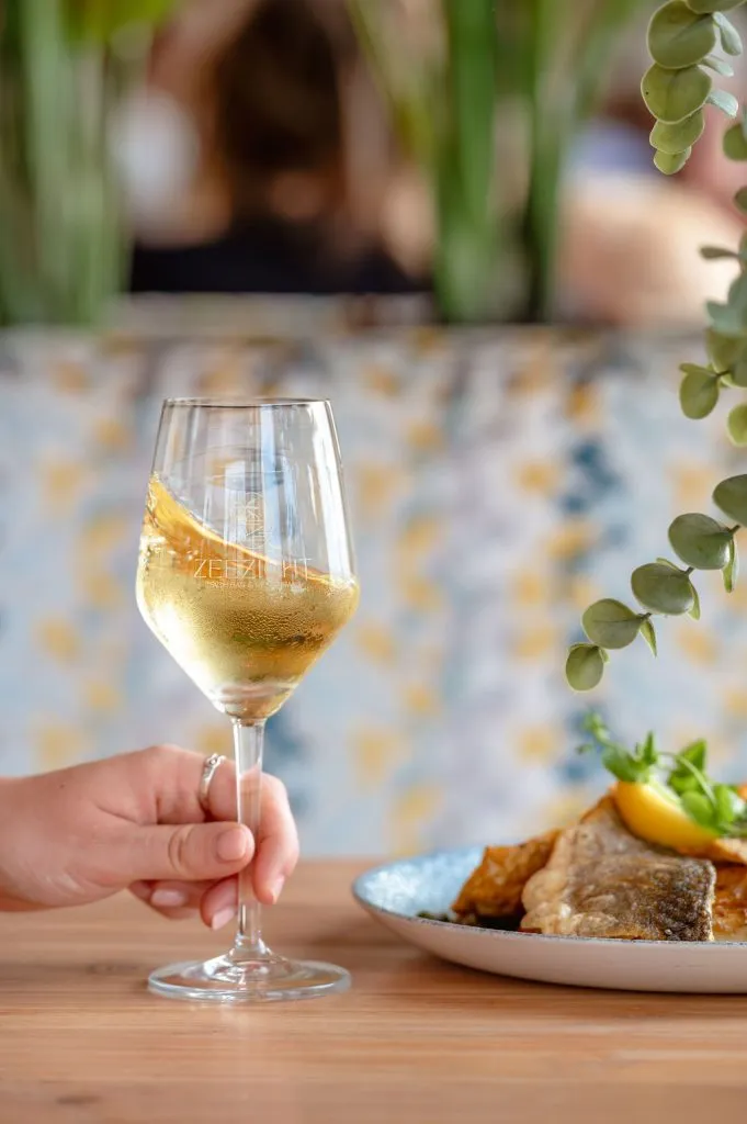 Close-up van een hand die een glas witte wijn vasthoudt met een schijfje sinaasappel, naast een vers visgerecht.