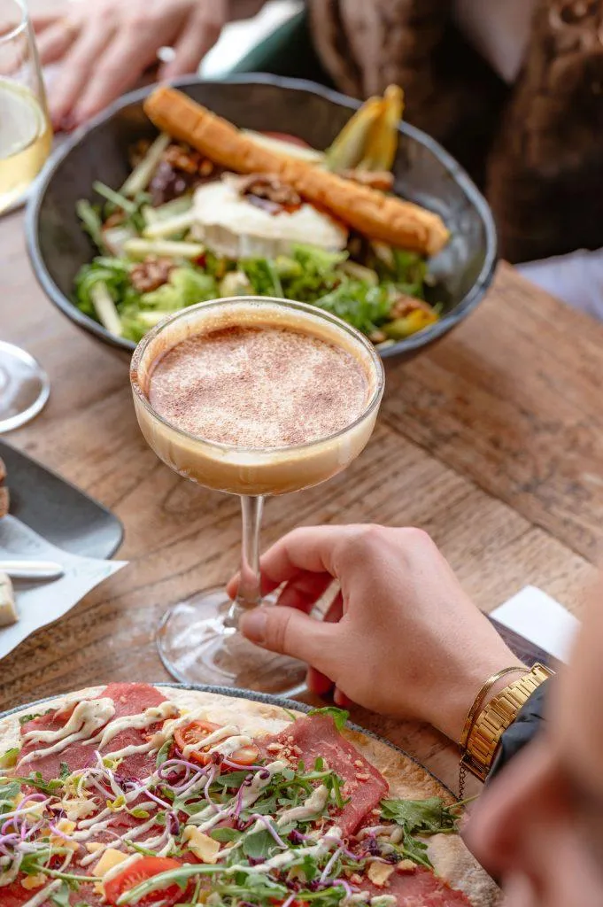 Espresso martini cocktail vastgehouden bij een tafel met een salade en carpaccio wrap.