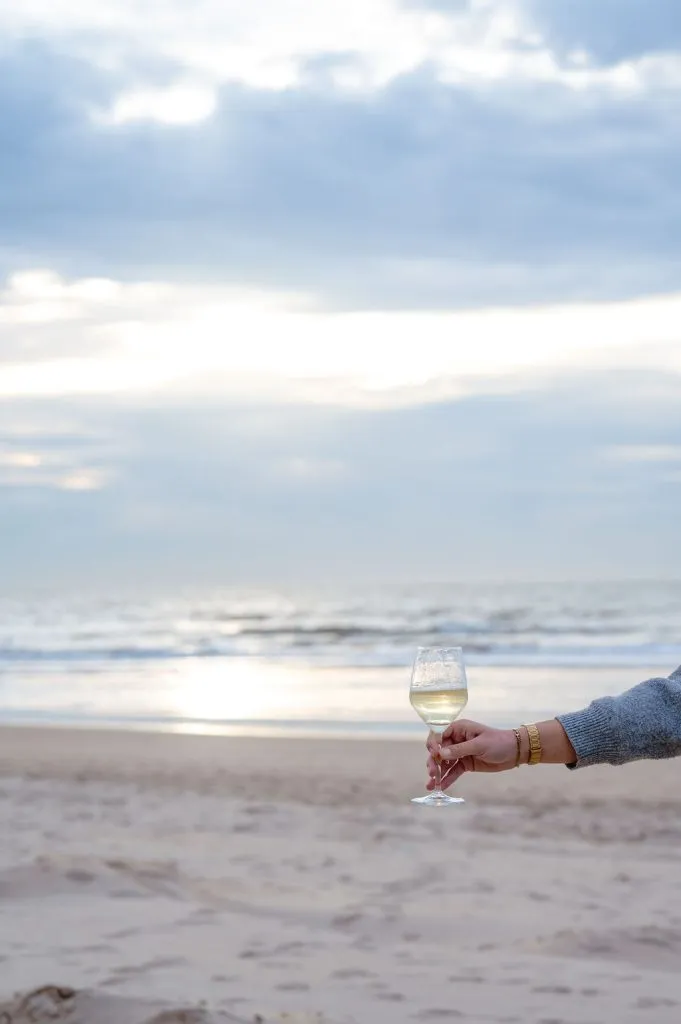 Een hand houdt een glas witte wijn vast op het strand met de Noordzee op de achtergrond bij zonsondergang.