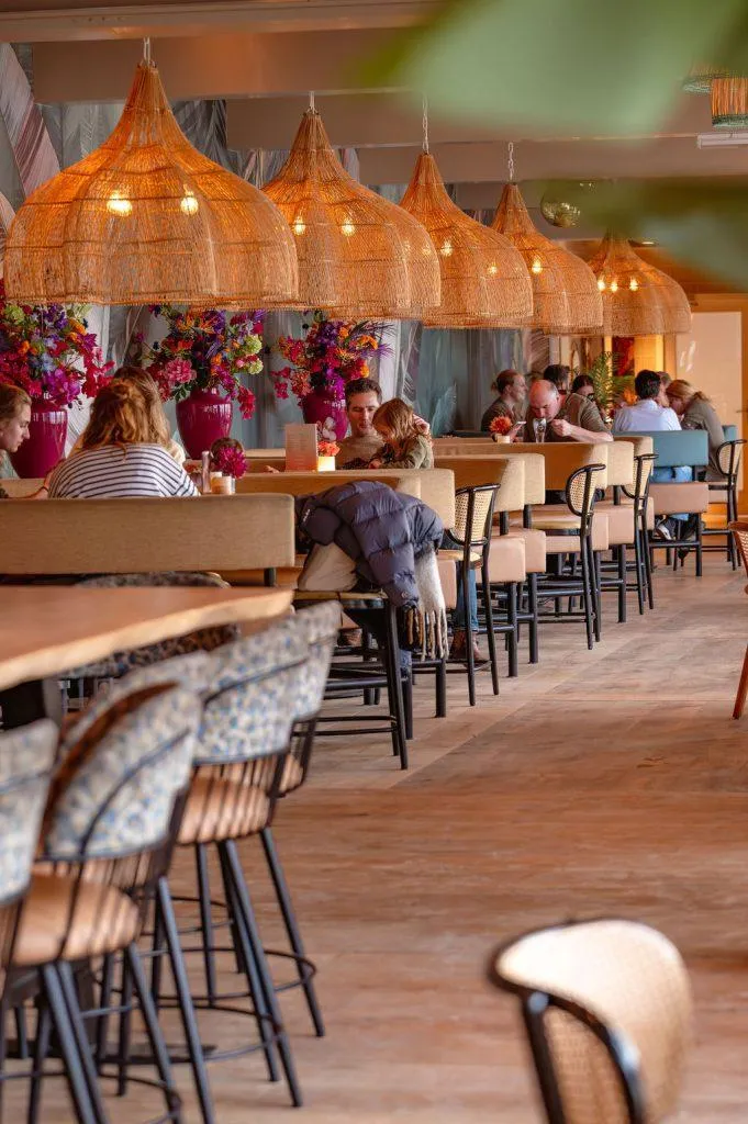 Gasten tafelen in een sfeervol interieur met grote rotan hanglampen en kleurrijke boeketten bloemen.