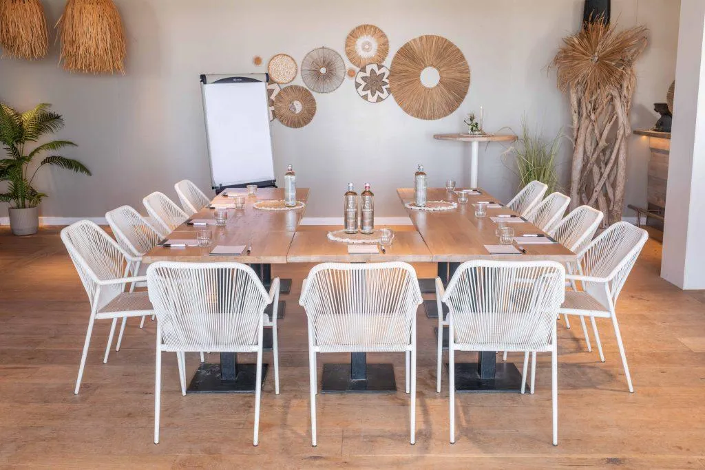 Vergaderopstelling aan zee met houten tafels, witte stoelen en sfeervolle stranddecoratie.