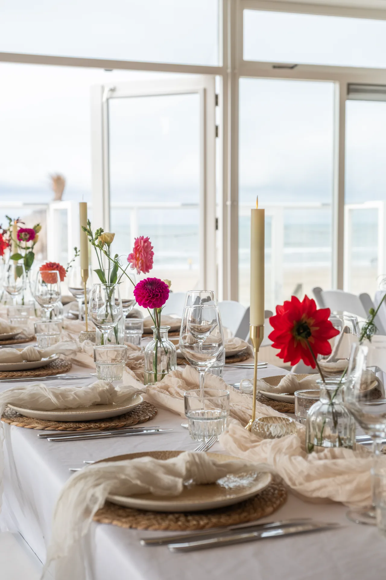 Elegant gedekte lange tafel aan zee met kleurrijke bloemen, kaarsen en linnen lopers.