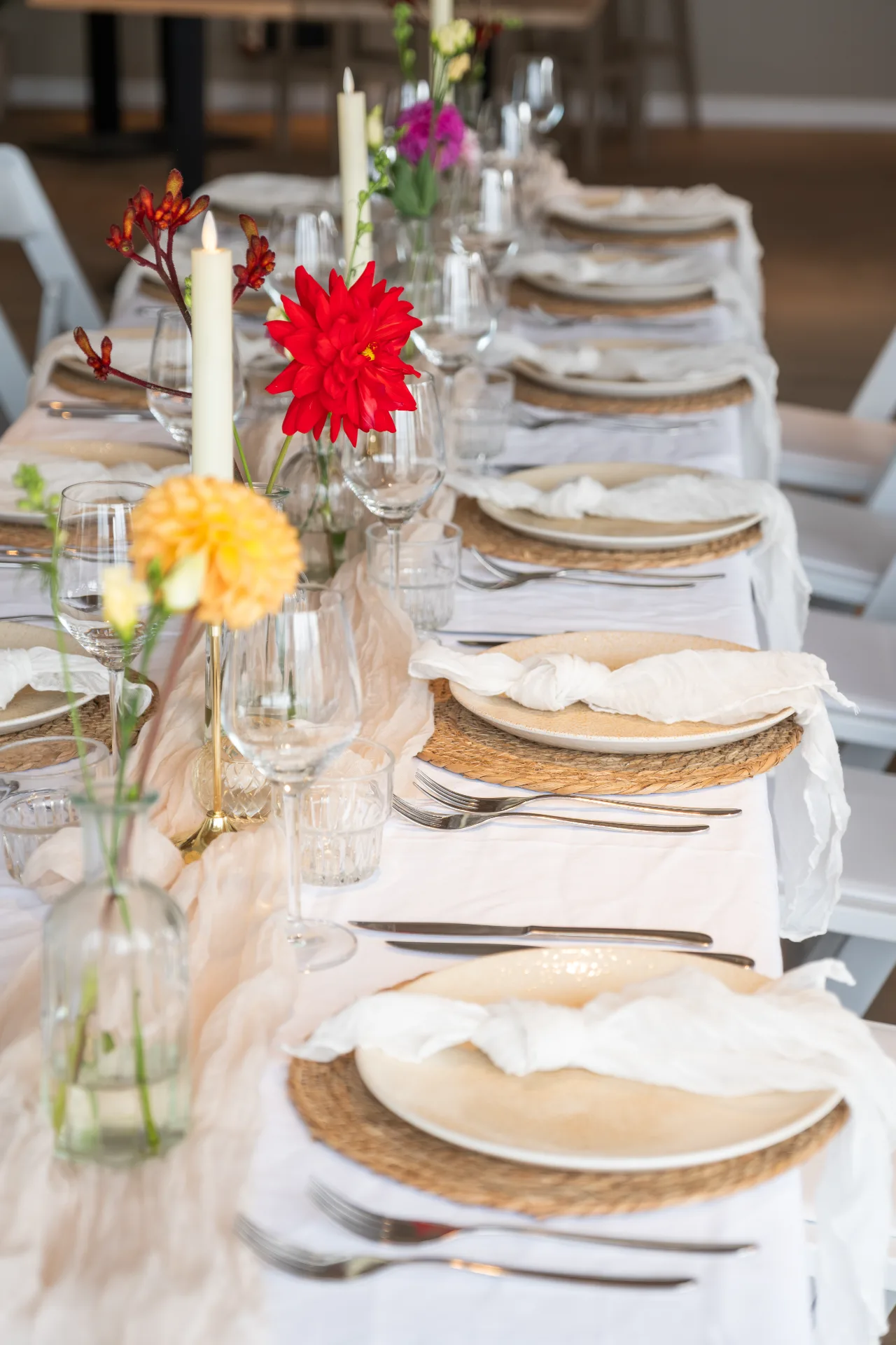 Lange gedekte dinertafel met wit linnen, kleurrijke bloemen, kaarsen en rustieke placemats.