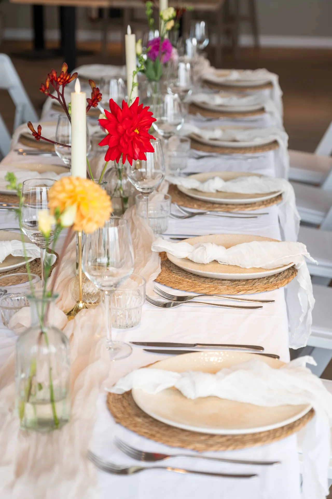 Lange gedekte dinertafel met wit linnen, kleurrijke bloemen, kaarsen en rustieke placemats in een strandpaviljoen.