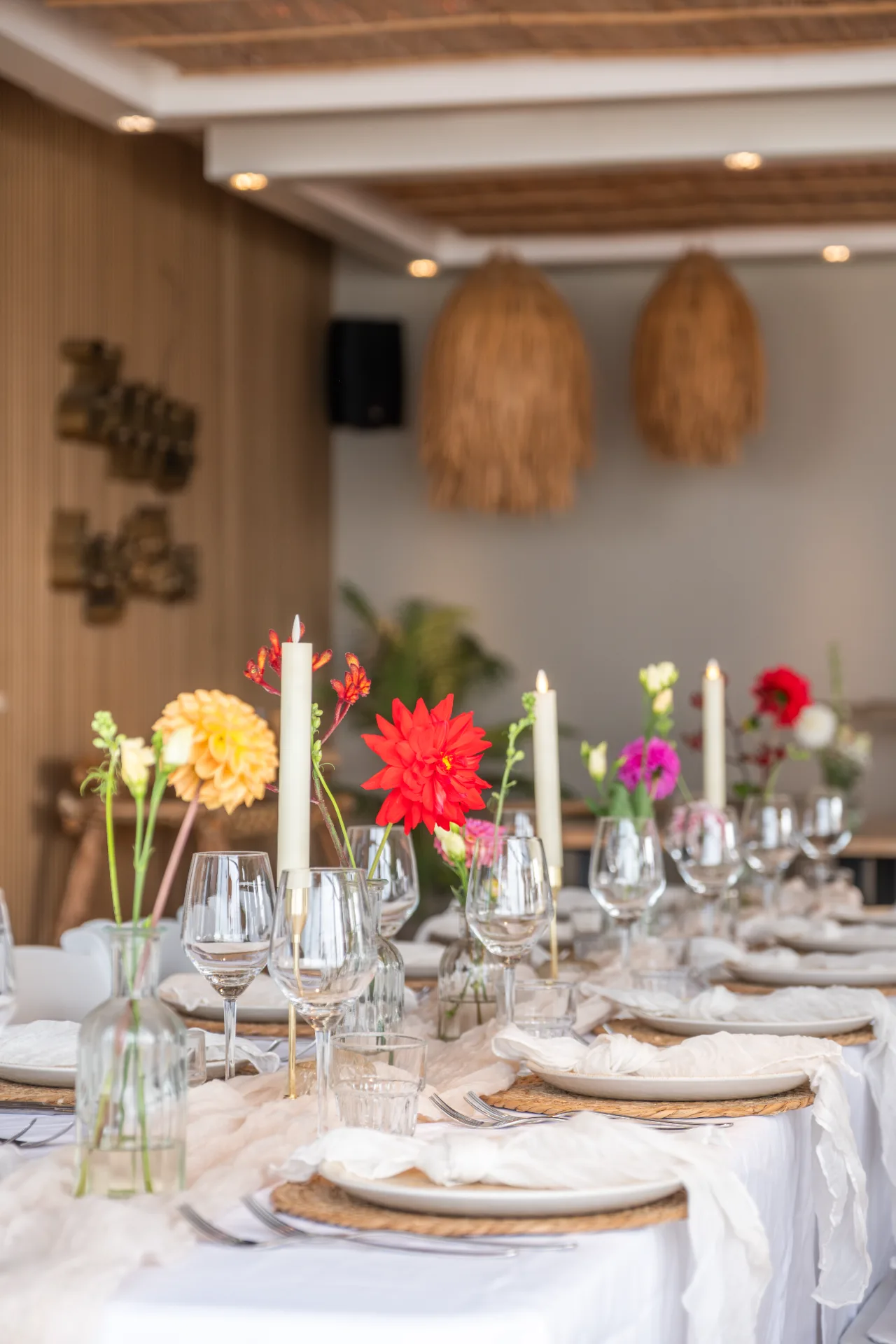Lange gedekte diner tafel met kleurrijke bloemen, witte kaarsen en wijnglazen in een strandpaviljoen.