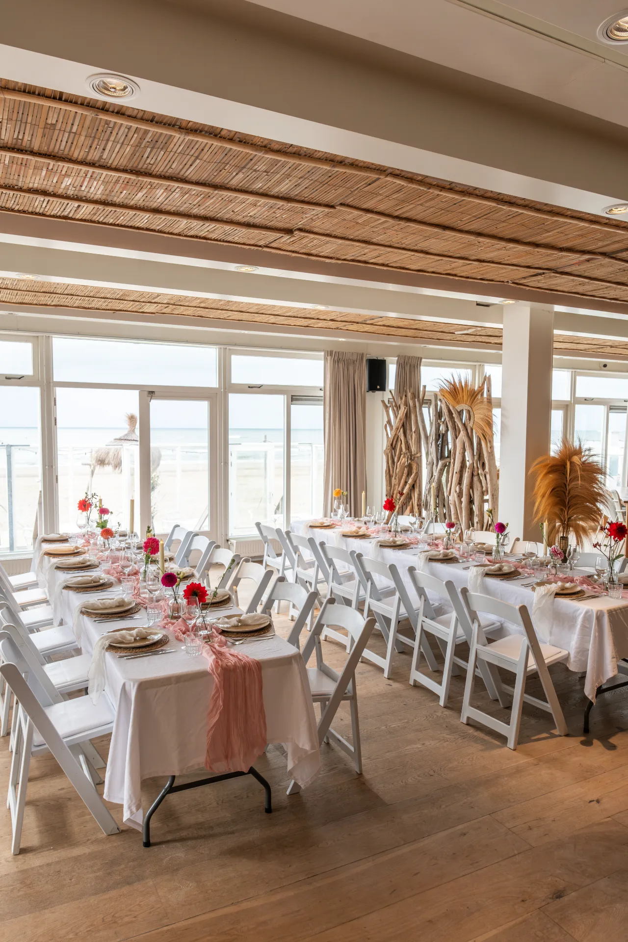 Gedekte lange tafels voor een diner met witte stoelen en roze lopers in een strandpaviljoen met zeezicht.