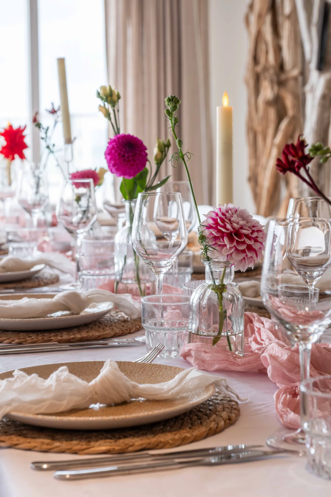 Sfeervol gedekte dinertafel met roze dahlia's, linnen servetten en wijnglazen bij een strandpaviljoen.